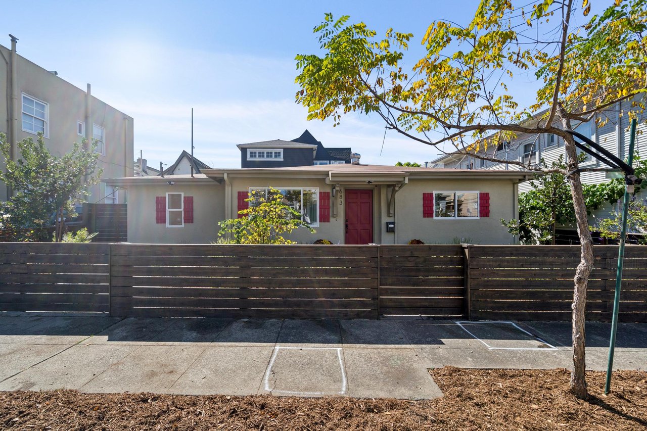 Gray single-story home with a red door and shutters behind a dark wood fence.