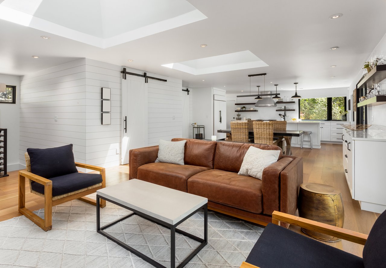 Open-plan living room with a brown leather sofa, navy chairs, and a bright kitchen beyond.