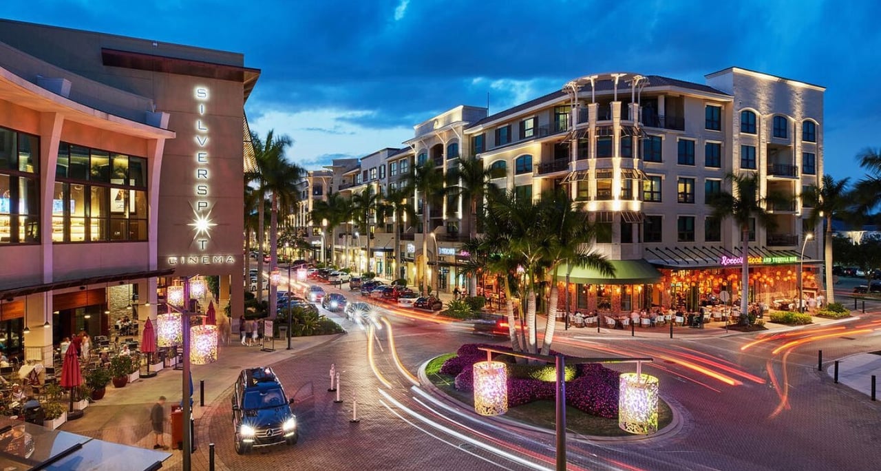 Illuminated shops, restaurants, and Silverspot Cinema line a palm-lined shopping plaza at dusk.