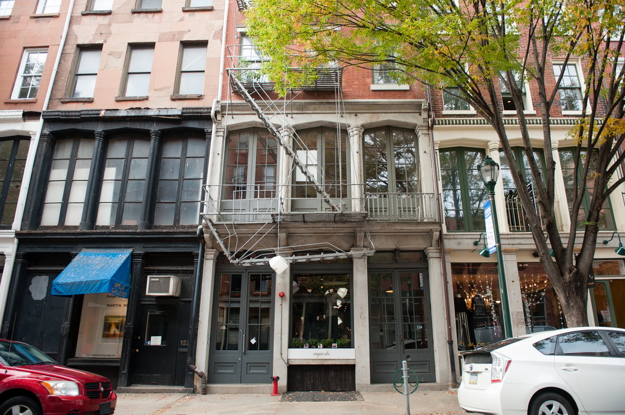 Historic attached buildings with storefronts, decorative columns, and a metal fire escape beneath a leafy street tree.