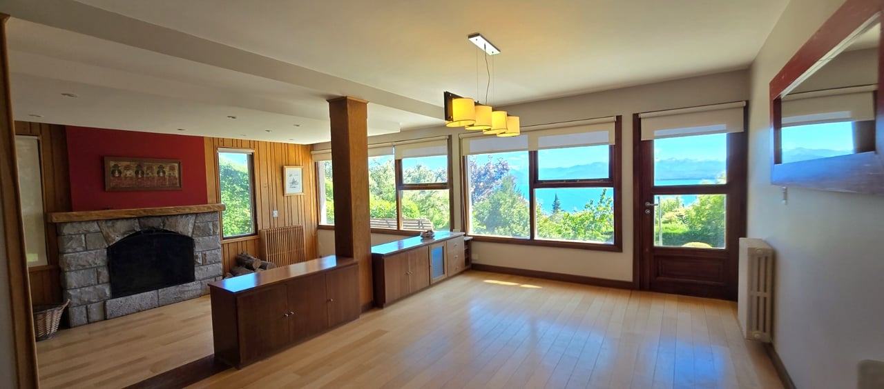 Living room with a stone fireplace and broad lake and mountain views through large windows.
