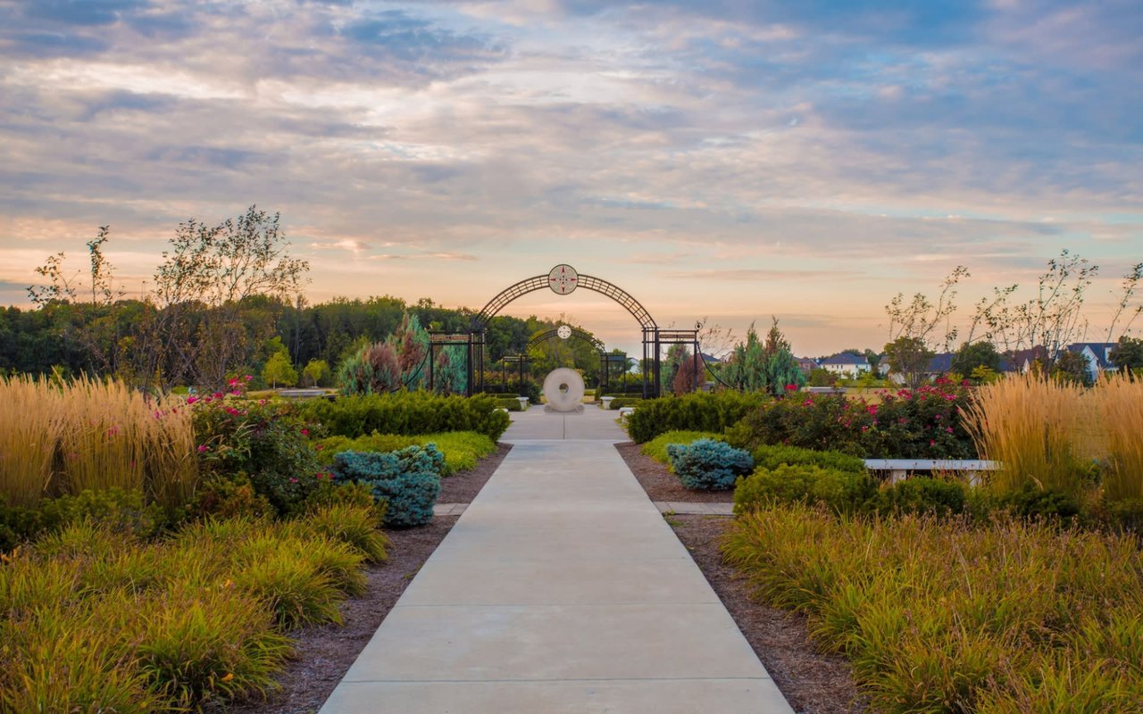 Beautifully landscaped public park walkway featuring structured garden beds, ornamental grasses, decorative metal trellis arches, and a sunset horizon at Coxhall Gardens.