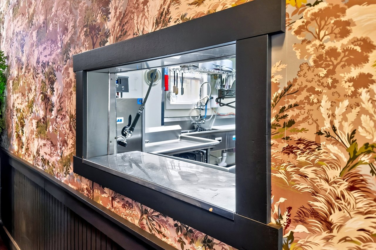 Restaurant service window reveals stainless-steel kitchen equipment, framed by ornate floral wallpaper.