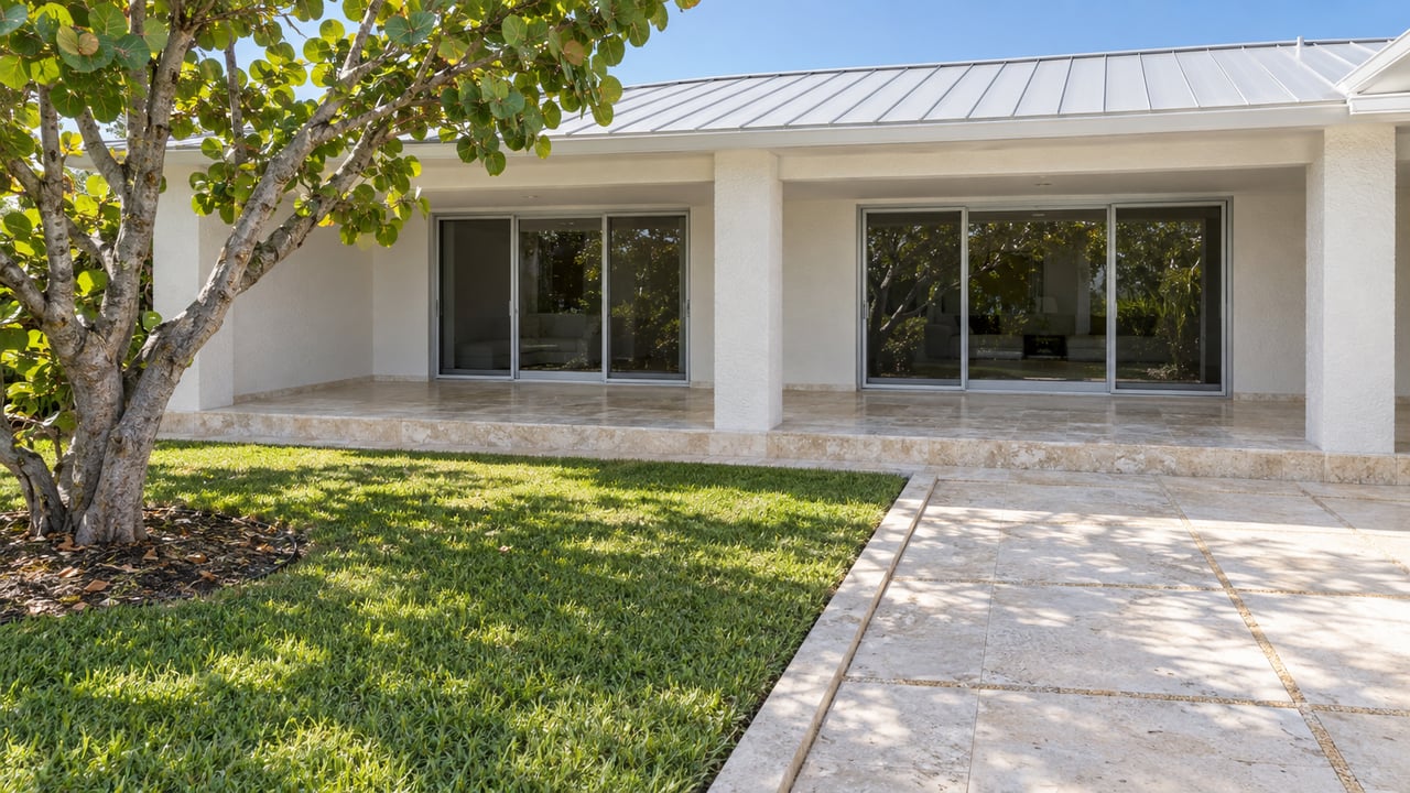 White stucco low-rise condominium with closed sliding-glass doors, pale stone terraces, and a mature sea grape tree beside a landscaped courtyard.