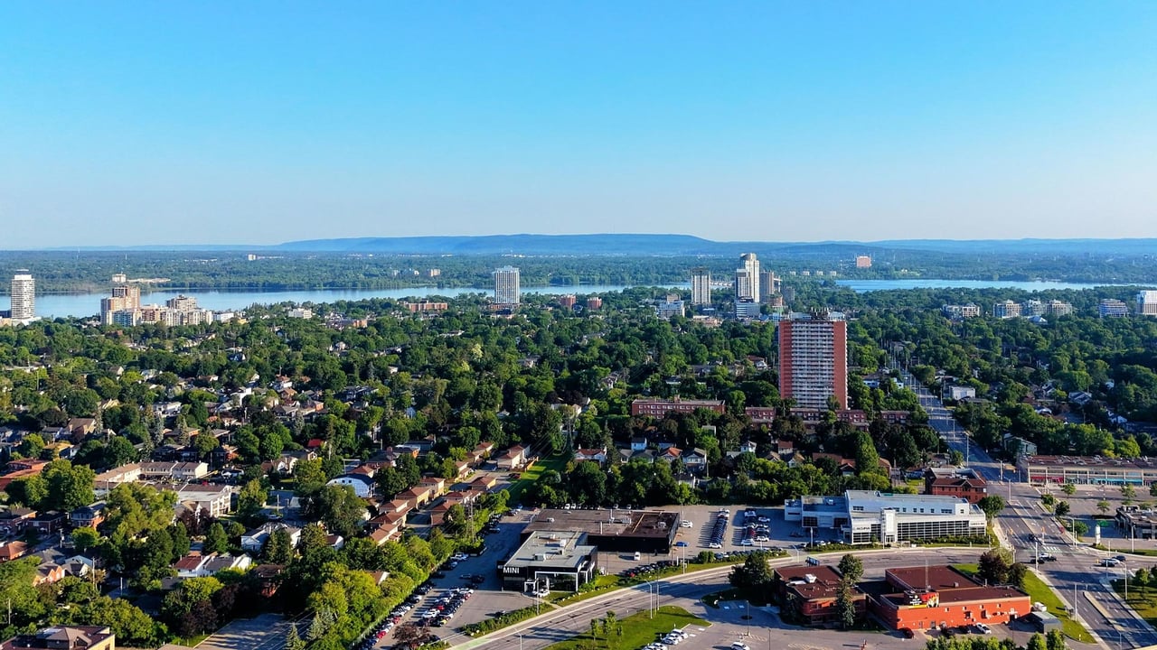 An expansive aerial view of Hamilton, Ontario showing vibrant green residential neighbourhoods, high-rise buildings, and the Hamilton Harbour with the Niagara Escarpment in the background, illustrating the lifestyle and location appeal for GTA home buyers.
