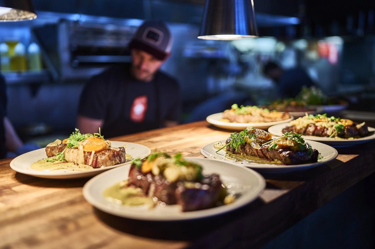 Plated steaks with herbs and sauce lined up on a wooden counter in a restaurant kitchen.