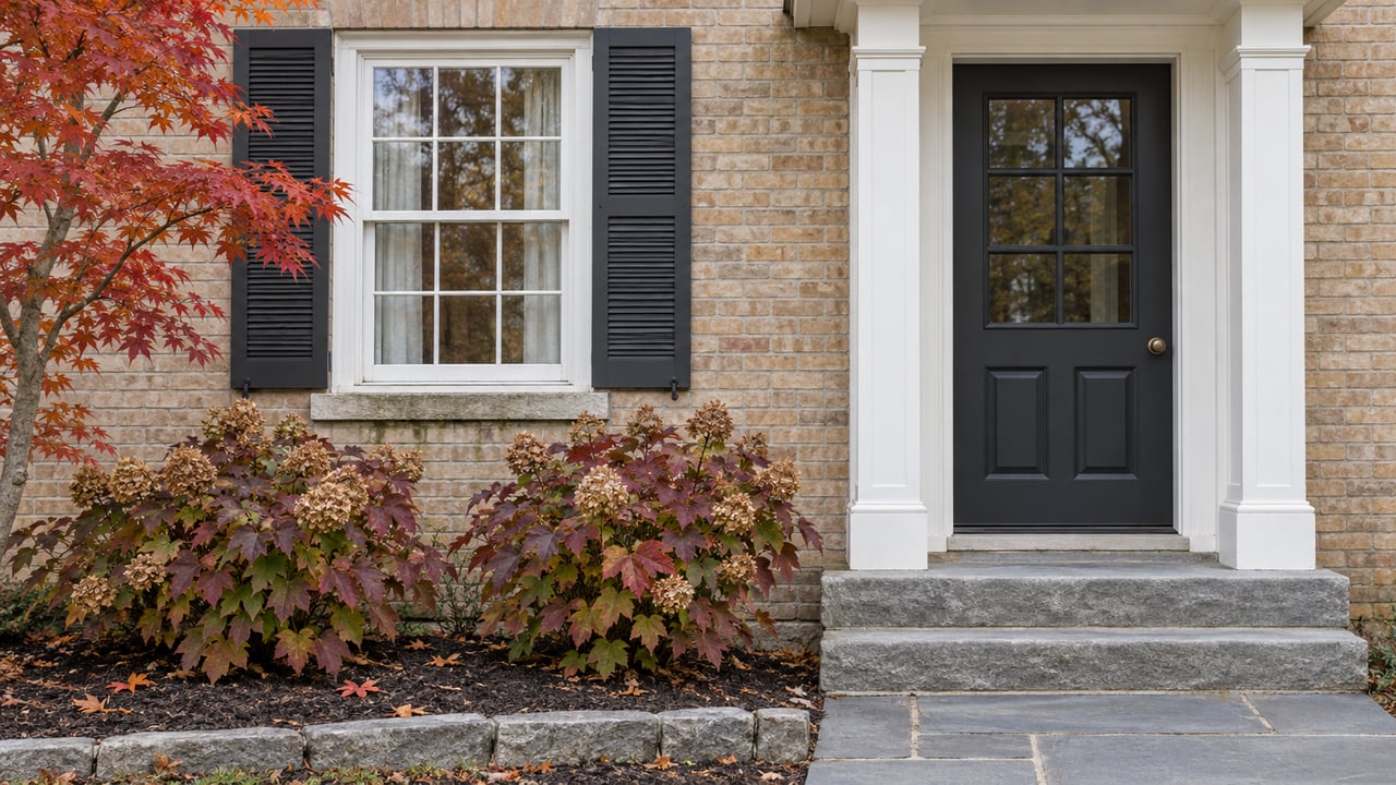 Pale brick home with a white-trimmed entrance, dark shutters, limestone steps, and burgundy autumn planting.
