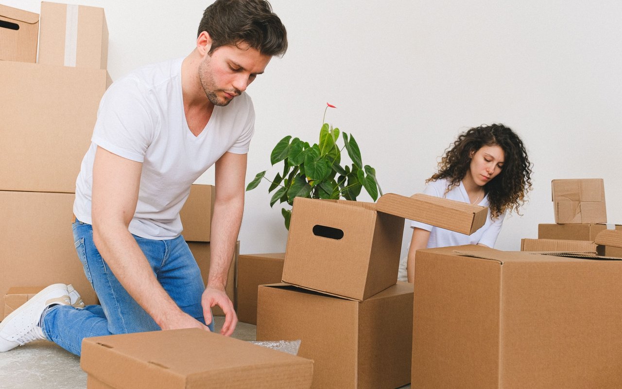 A man and woman pack cardboard boxes in a mostly empty room.