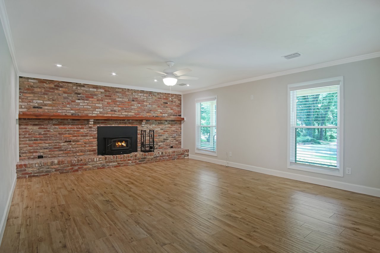 Spacious living room with a rustic brick fireplace, wooden mantel, and light wood floors. Large windows and neutral walls create a bright, airy feel.