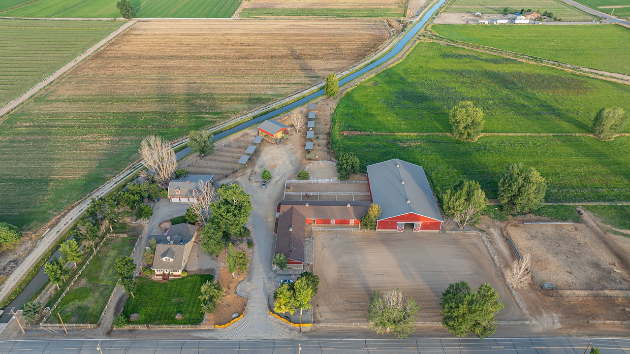 Aerial ranch with red barns, a riding arena, a farmhouse, and irrigated fields beside a canal.