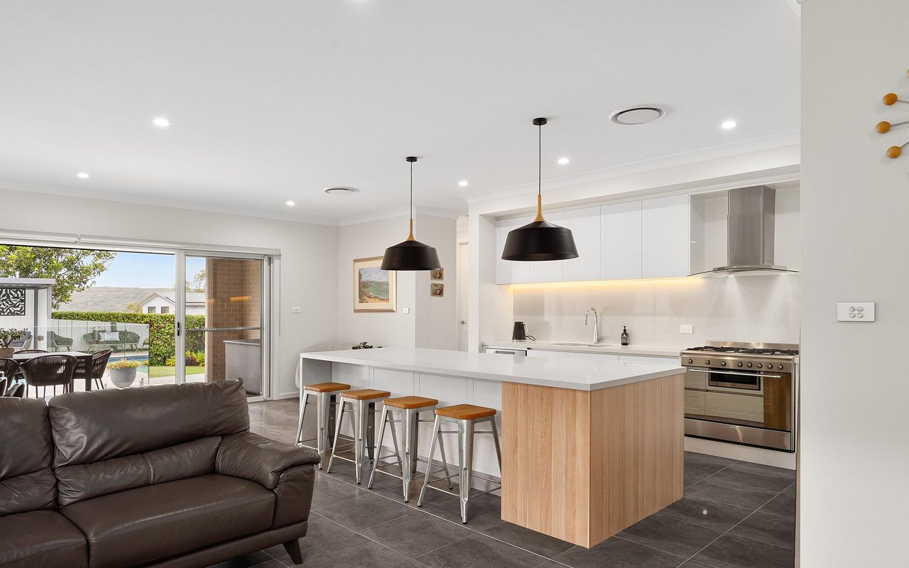 Open-plan kitchen with a wood-front island, dark leather sofa, and sliding doors overlooking a backyard pool.