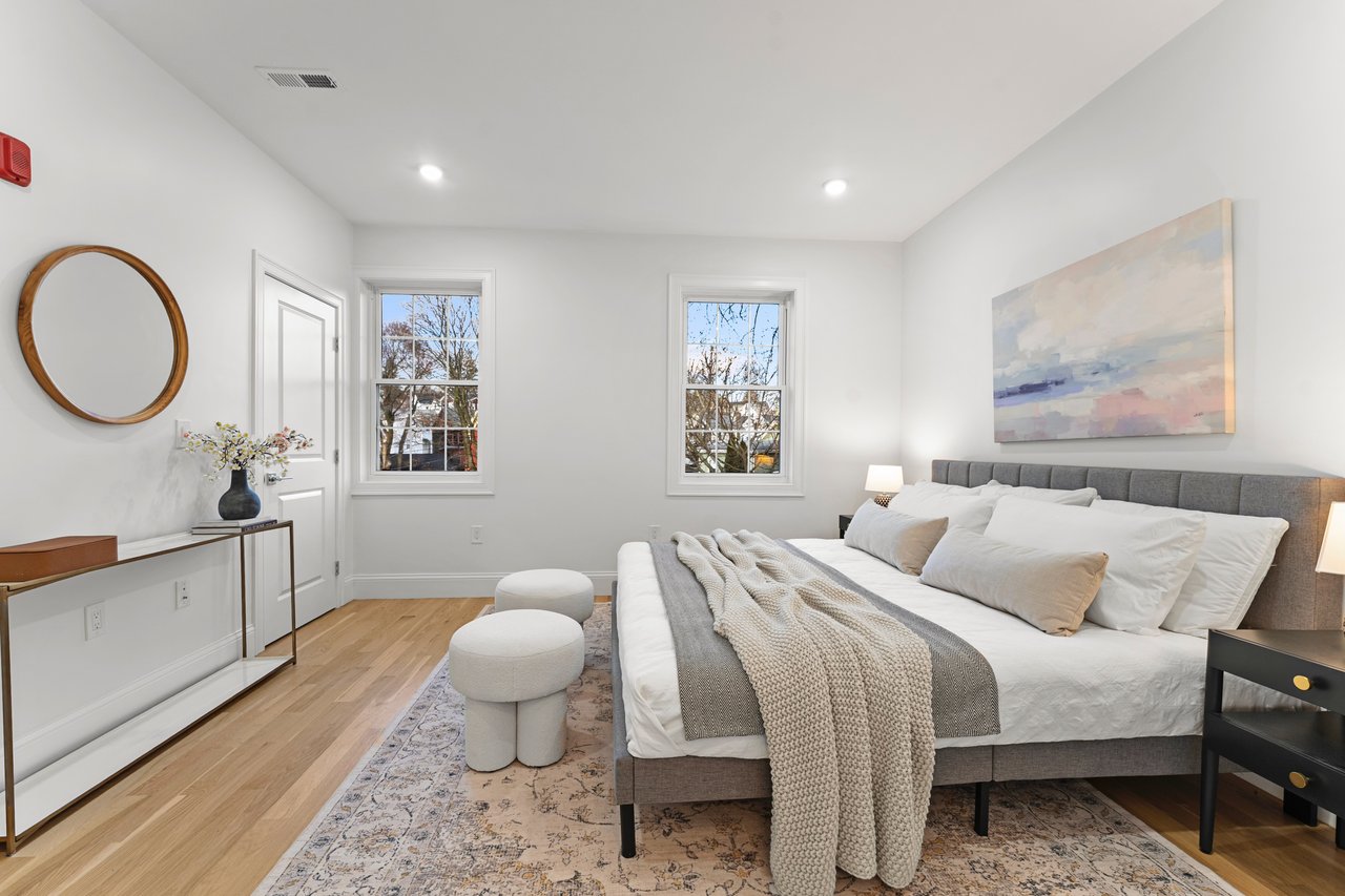Staged bedroom with a gray upholstered bed, white bedding, two windows, and light wood floors.