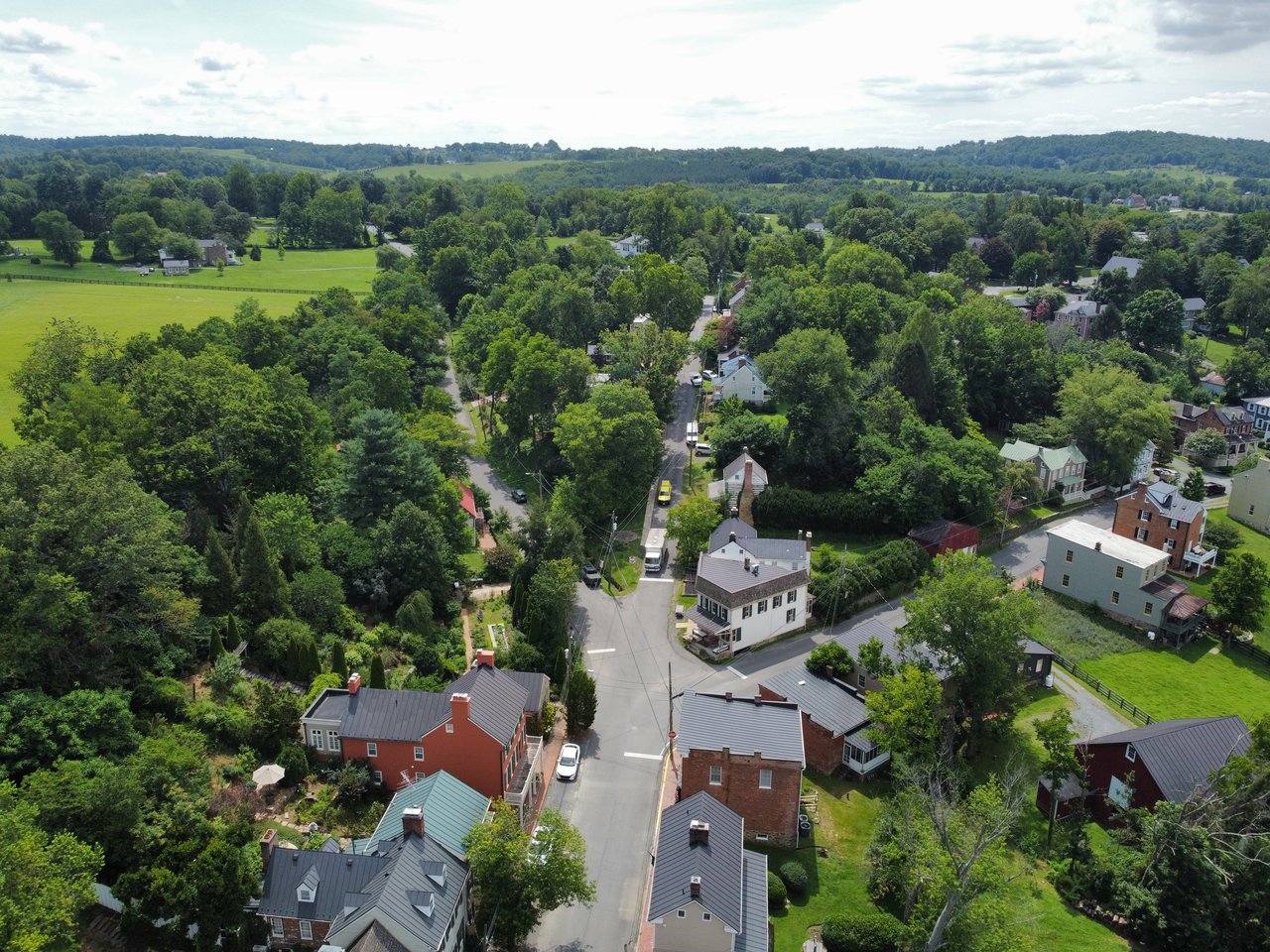 A sweeping high-altitude aerial drone shot showcasing the historic stone buildings, rolling green farmland, and countryside landscape of Waterford, VA, captured by local real estate expert Diana Geremia.  Live On-Page Caption: A bird's-eye perspective of the rolling countryside and historic architecture that define Waterford, VA! — Contributed by local real estate expert Diana Geremia.