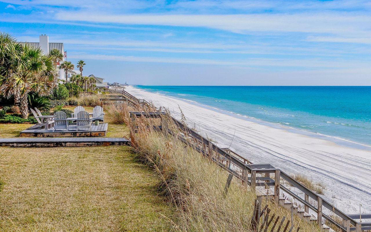 White-sand beach and turquoise water beside beachfront homes, dune grass, and a wooden boardwalk.