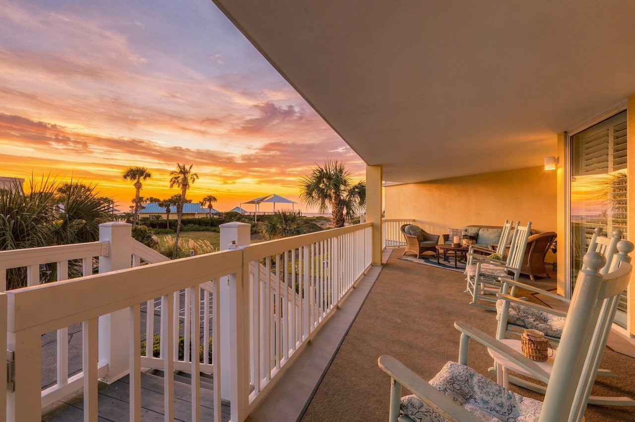 Covered balcony with outdoor seating overlooking palm trees and coastal dunes at sunset.