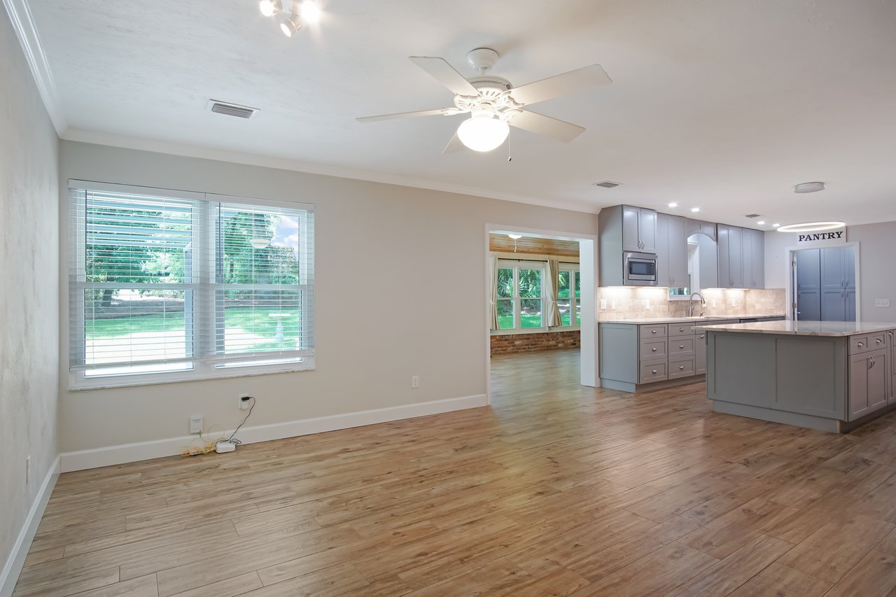 Spacious, modern kitchen with gray cabinets, a central island, and warm wooden flooring. Large windows offer natural light and a view of greenery.