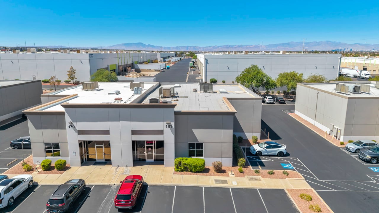 Commercial building with gray facade and storefronts, viewed above its parking lot with mountains in the distance.