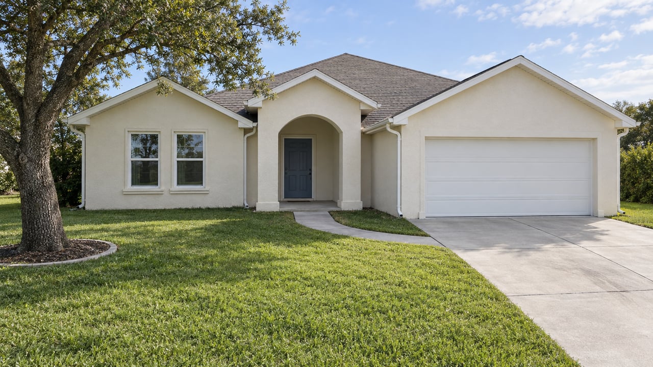 Cream stucco home with three gables, an arched entry, white garage door, deep front lawn and one mature live oak in bright late-morning light.