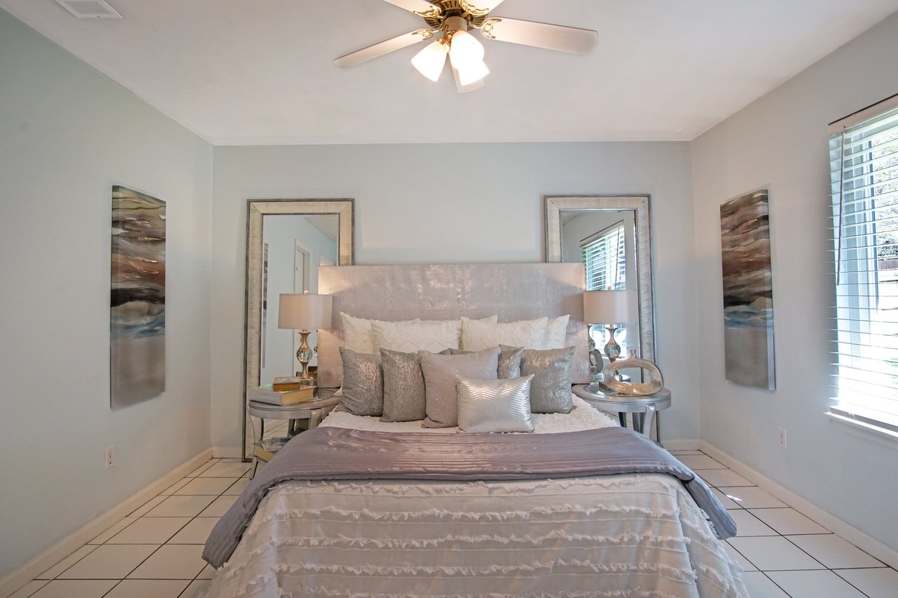 Elegant bedroom with a plush silver headboard, layered pillows, and a ruffled bedspread. Two mirrors and abstract art add a modern touch. Soft lighting creates a serene ambiance.