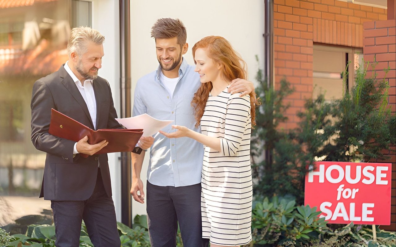 Real estate agent reviews paperwork with a couple beside a brick house and a for-sale sign.