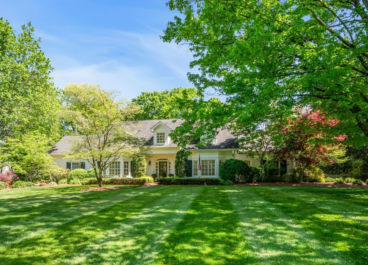 Large light-colored house framed by mature trees, with a broad, neatly mowed lawn.