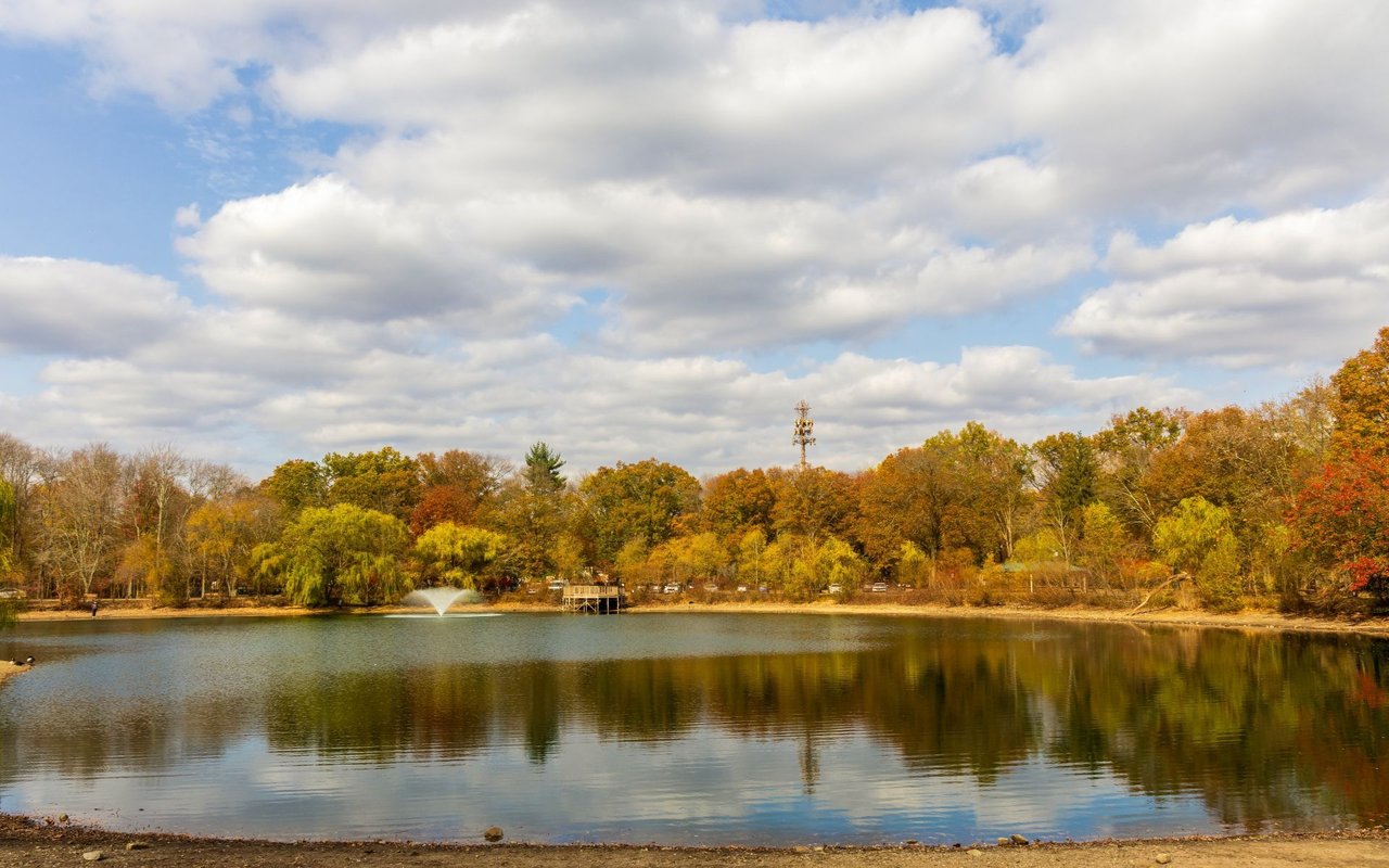 Pond with a fountain and dock, bordered by autumn trees and reflected in the water.