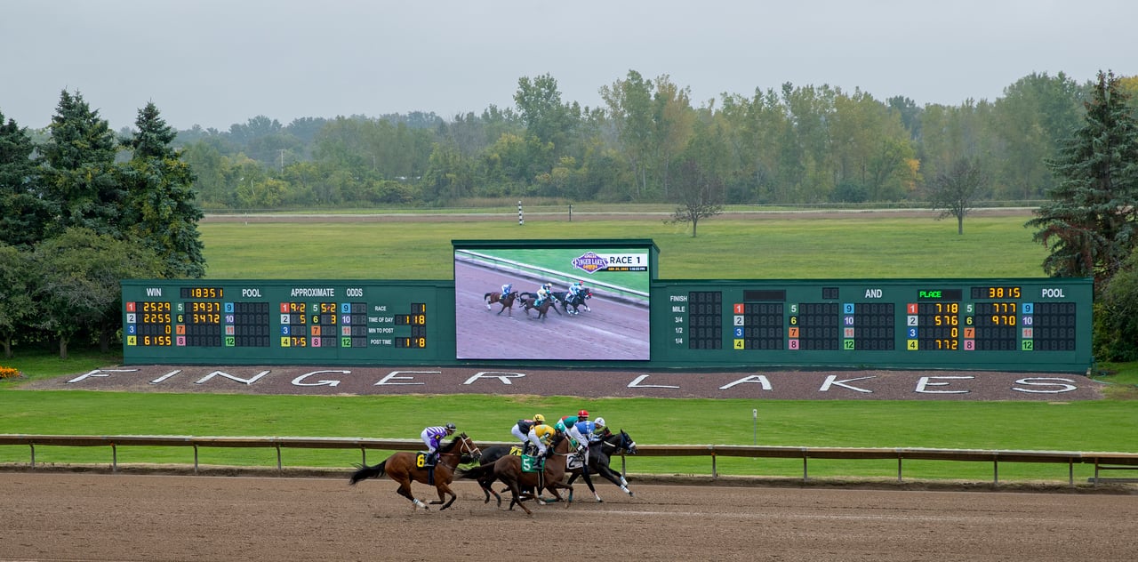 Horse race at Finger Lakes, with jockeys rounding the dirt track beneath the race results board.