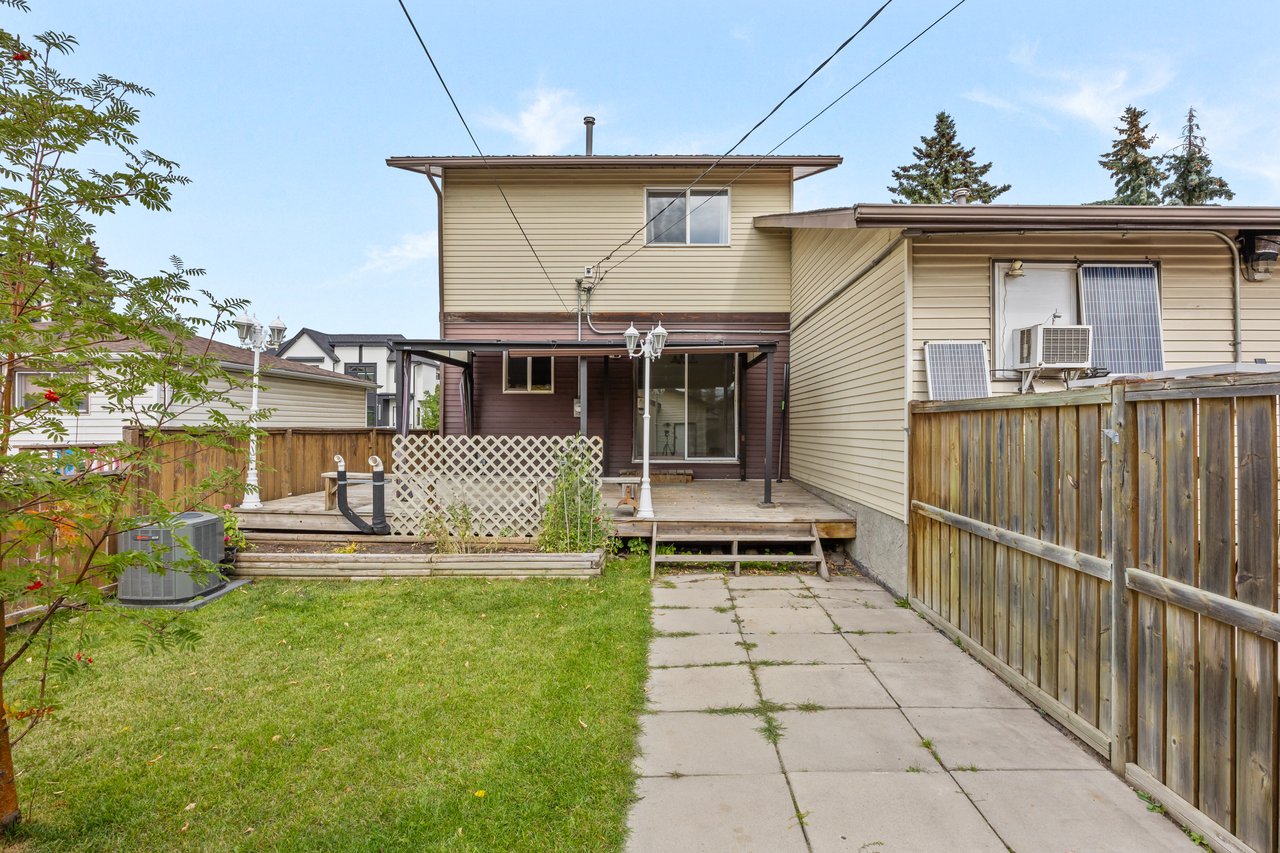 Rear of a beige two-story home with a raised deck, fenced lawn, and paved patio.