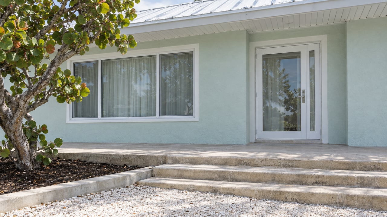 Seafoam home's glazed entry and white-trimmed window above worn concrete steps, beside a sea-grape tree.