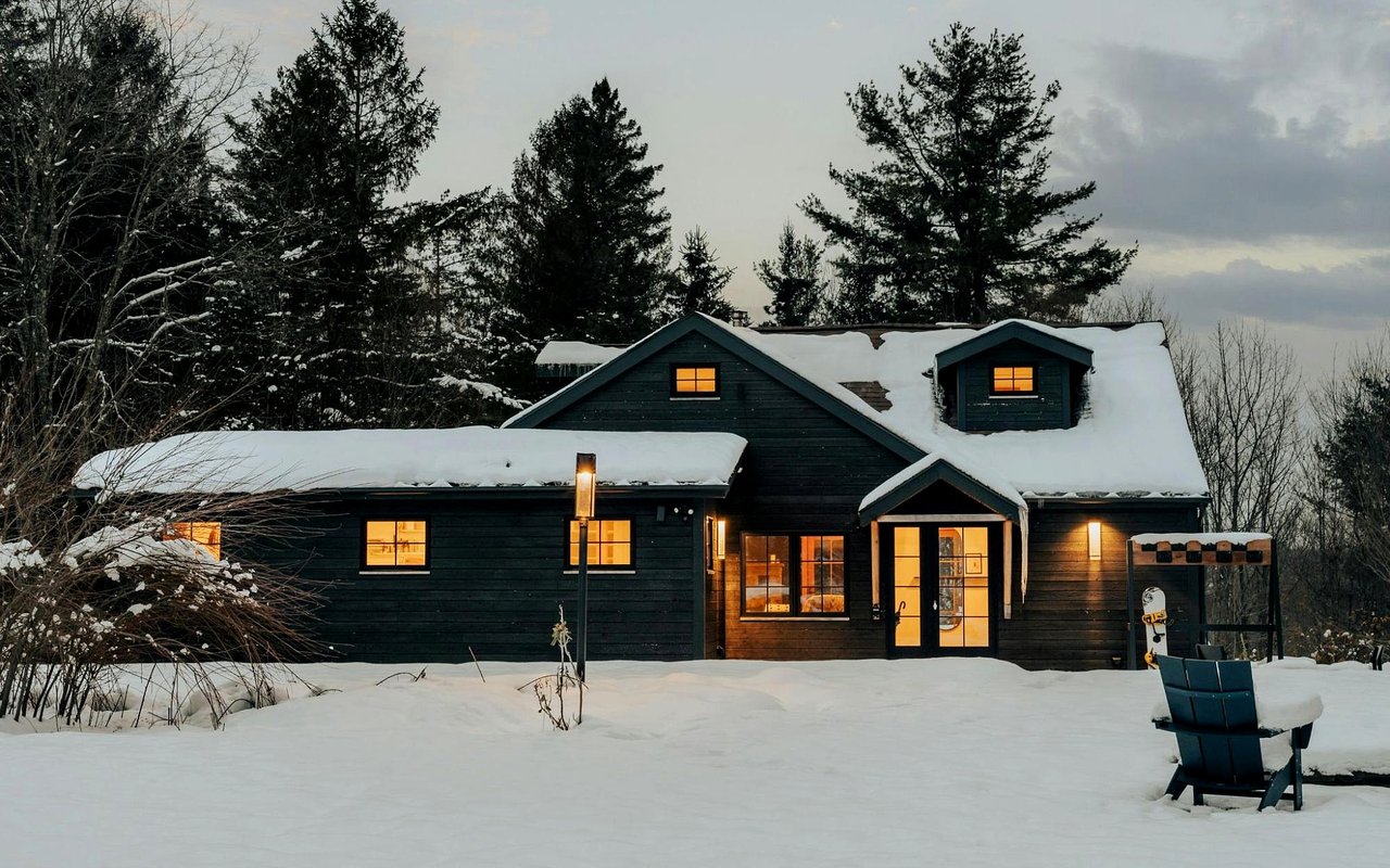 Dark wood house with glowing windows and snow-covered roof at dusk.