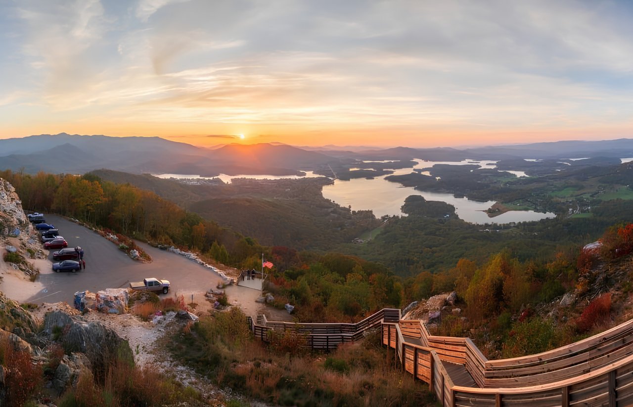 Winding lake and wooded mountain ridges at sunset, viewed from a rocky overlook with a wooden walkway.