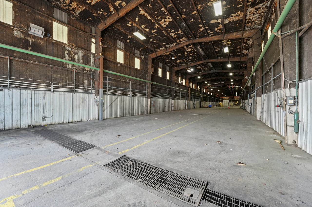 Vacant warehouse interior with a high exposed roof and long drainage grates across the concrete floor.