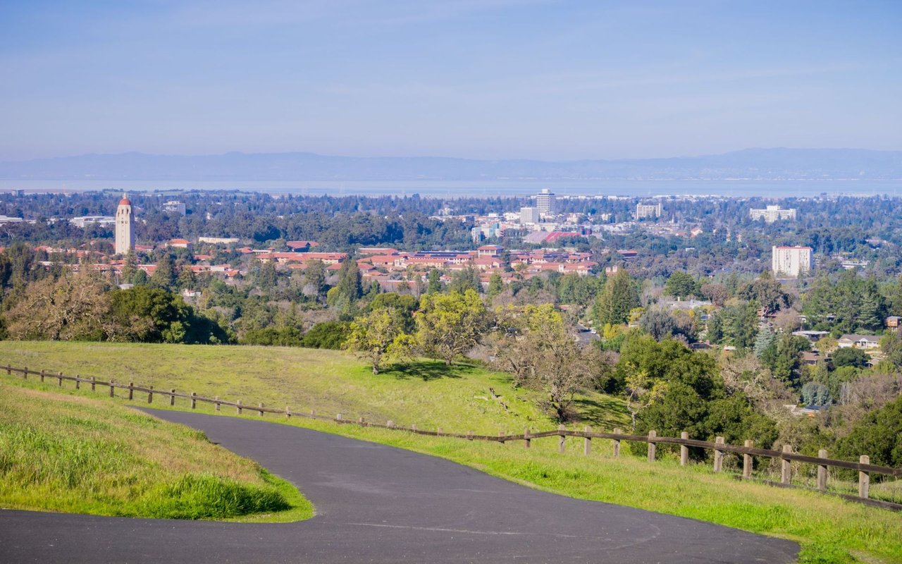 Panoramic campus view from a grassy hillside, with a prominent tower and distant water.