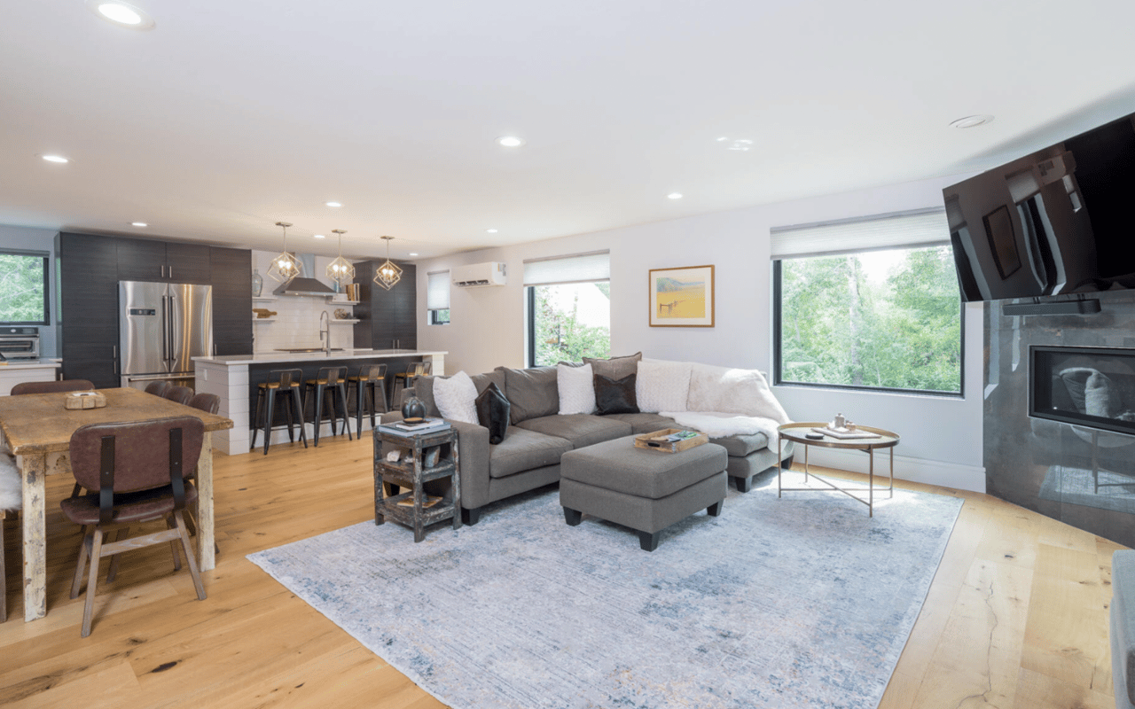 Open-plan living room with a gray sectional, light wood floors, and large windows overlooking trees.