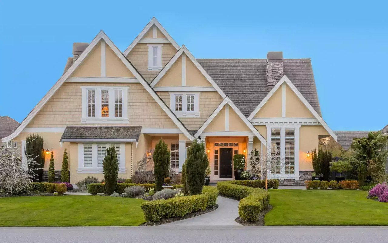 Beige shingle house with steep gables, landscaped hedges, and a curved front walkway.