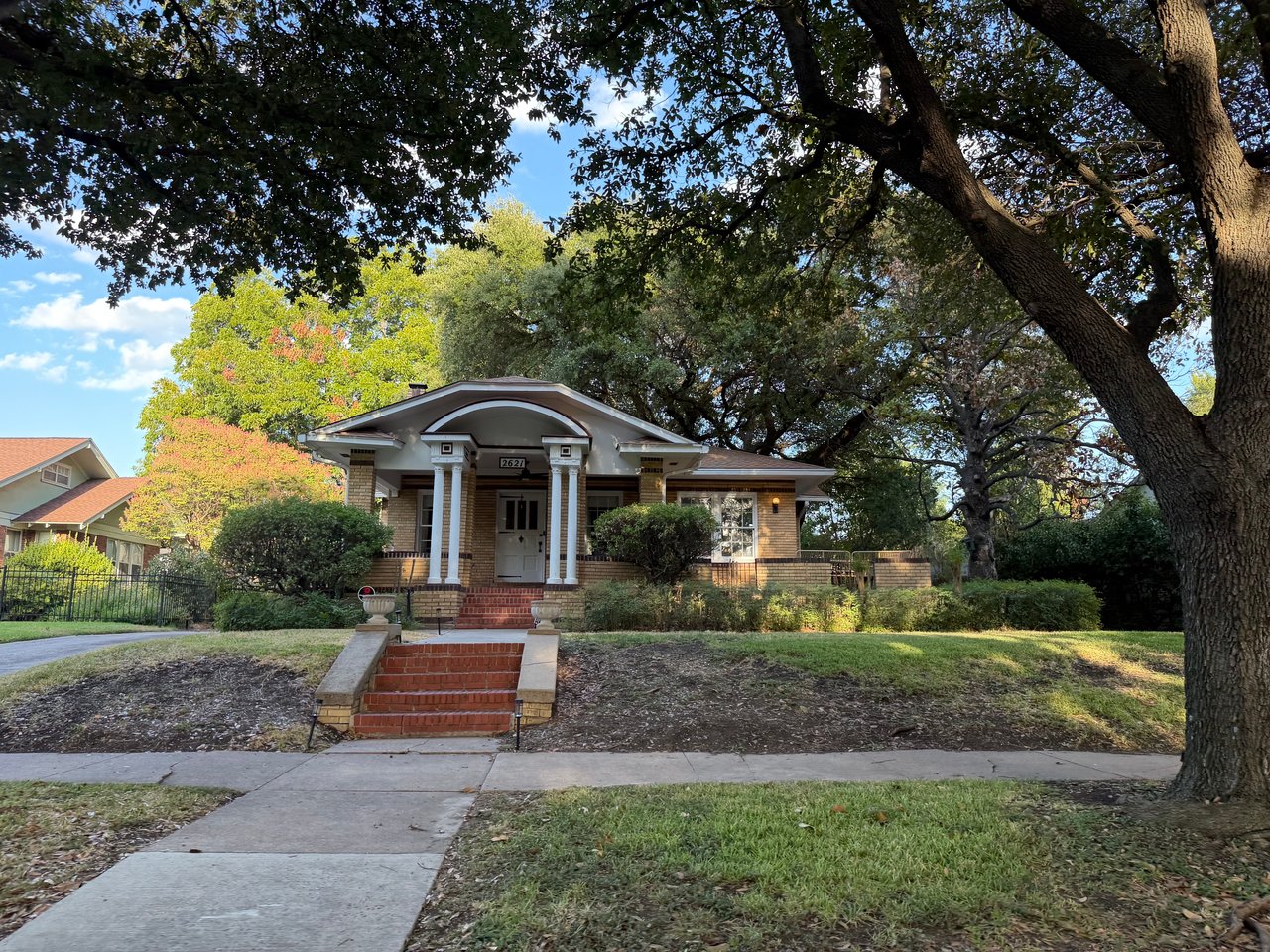 Brick home with an arched, columned porch and red brick steps beneath mature trees.