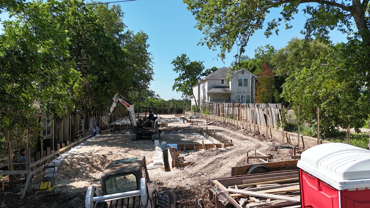 Residential development site under construction, with excavation equipment and foundation forms among mature trees.
