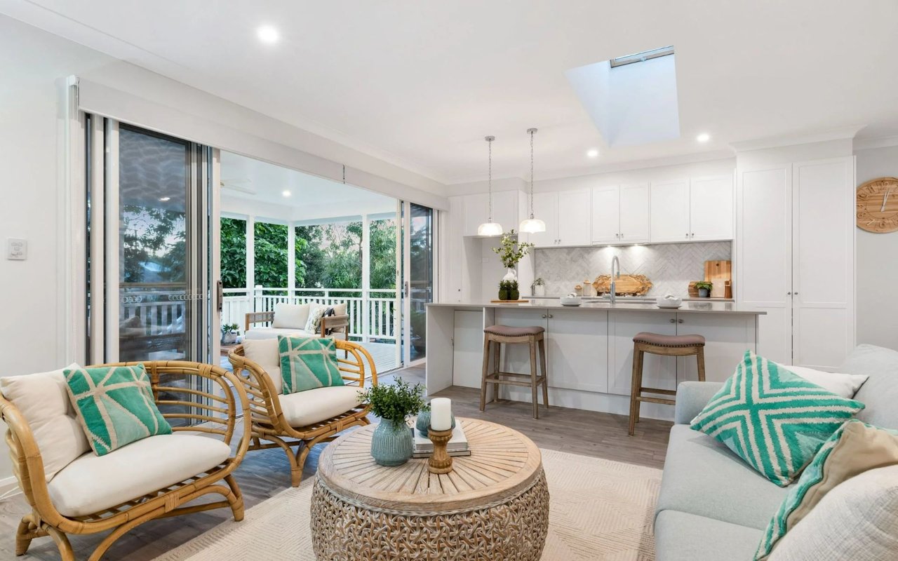 Open-plan living room and kitchen with rattan seating, turquoise pillows, and sliding doors to a leafy deck.
