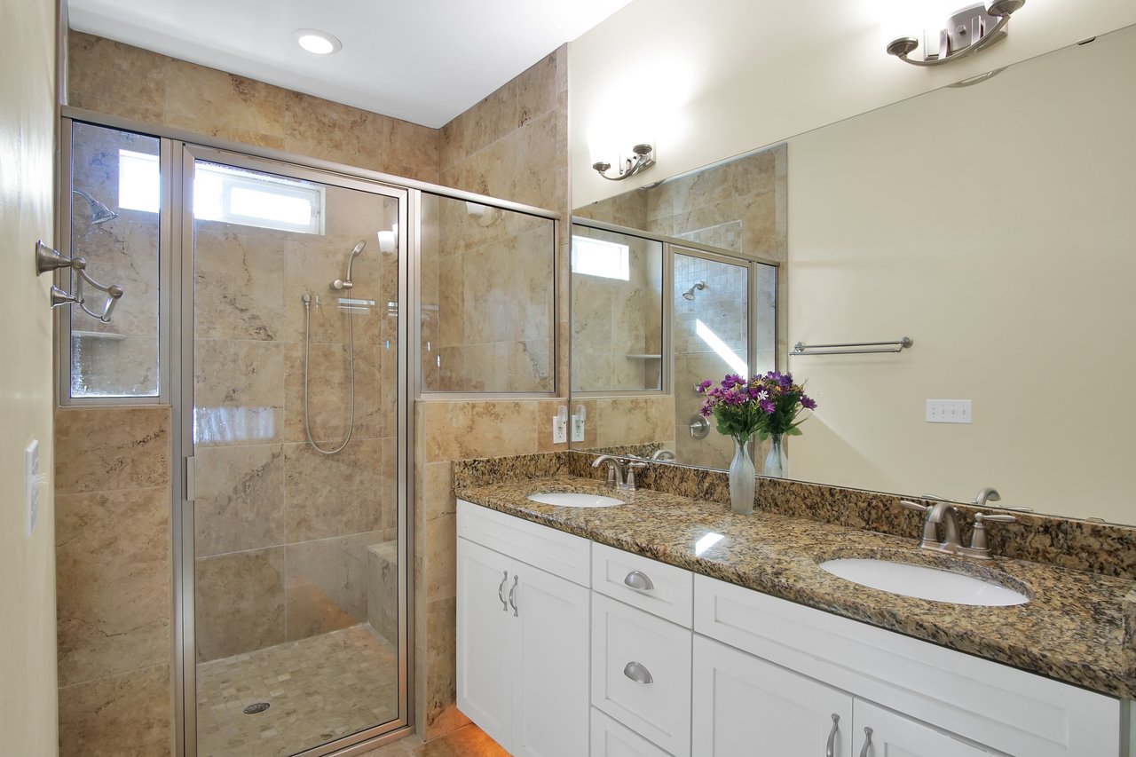 Elegant bathroom with marble tile shower, glass door, dual sink vanity with brown granite countertop, and a vase of purple flowers. Bright and inviting.