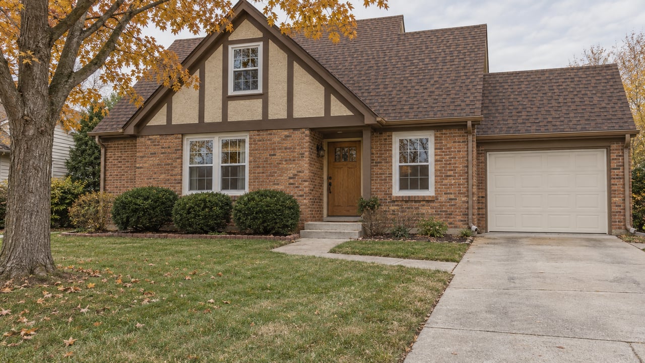 An orange-red brick and tan timber-trimmed home with a brown shingle roof, concrete front walk and an amber-leaved maple beside the lawn.