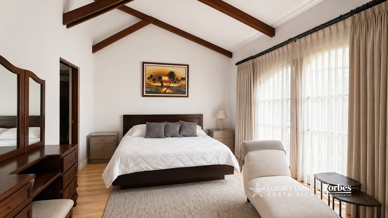 Bedroom with a dark-wood bed beneath exposed ceiling beams and tall curtained windows.