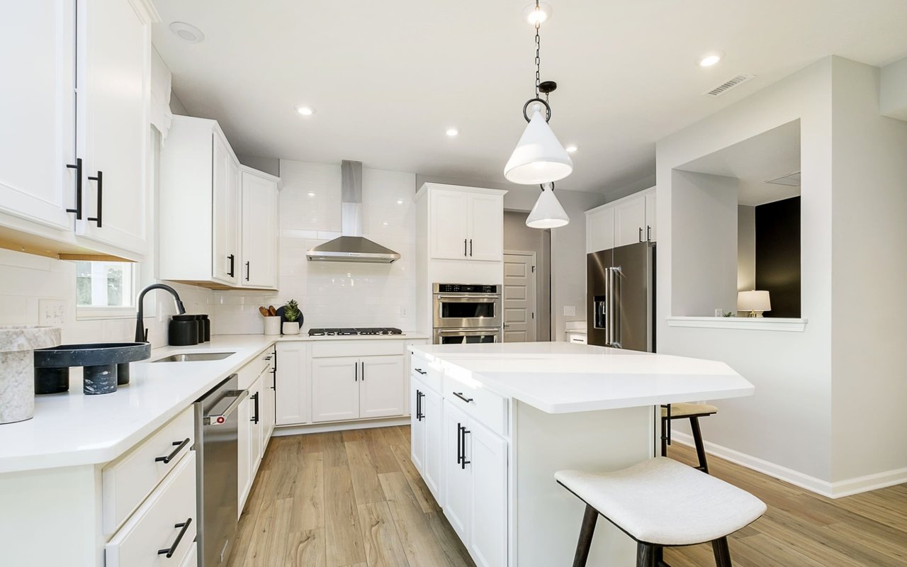 A bright, contemporary kitchen with white cabinets, black hardware handles, and white quartz countertops. The space features a large central island with bar stools, cone-shaped pendant lights, a stainless steel range hood, double ovens, and light-colored hardwood laminate flooring.