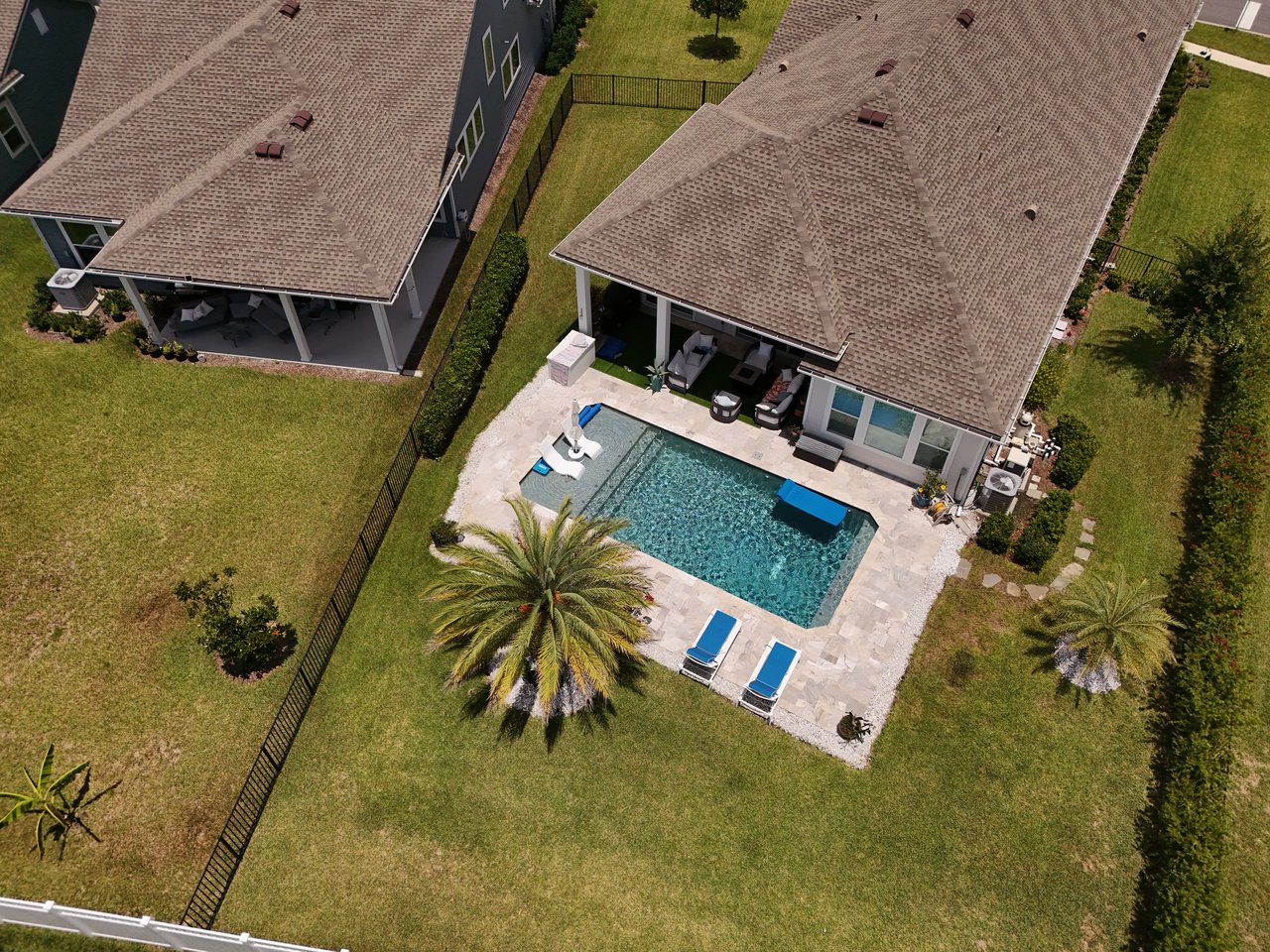 Aerial view of a home with a backyard pool, stone patio, and fenced lawn.