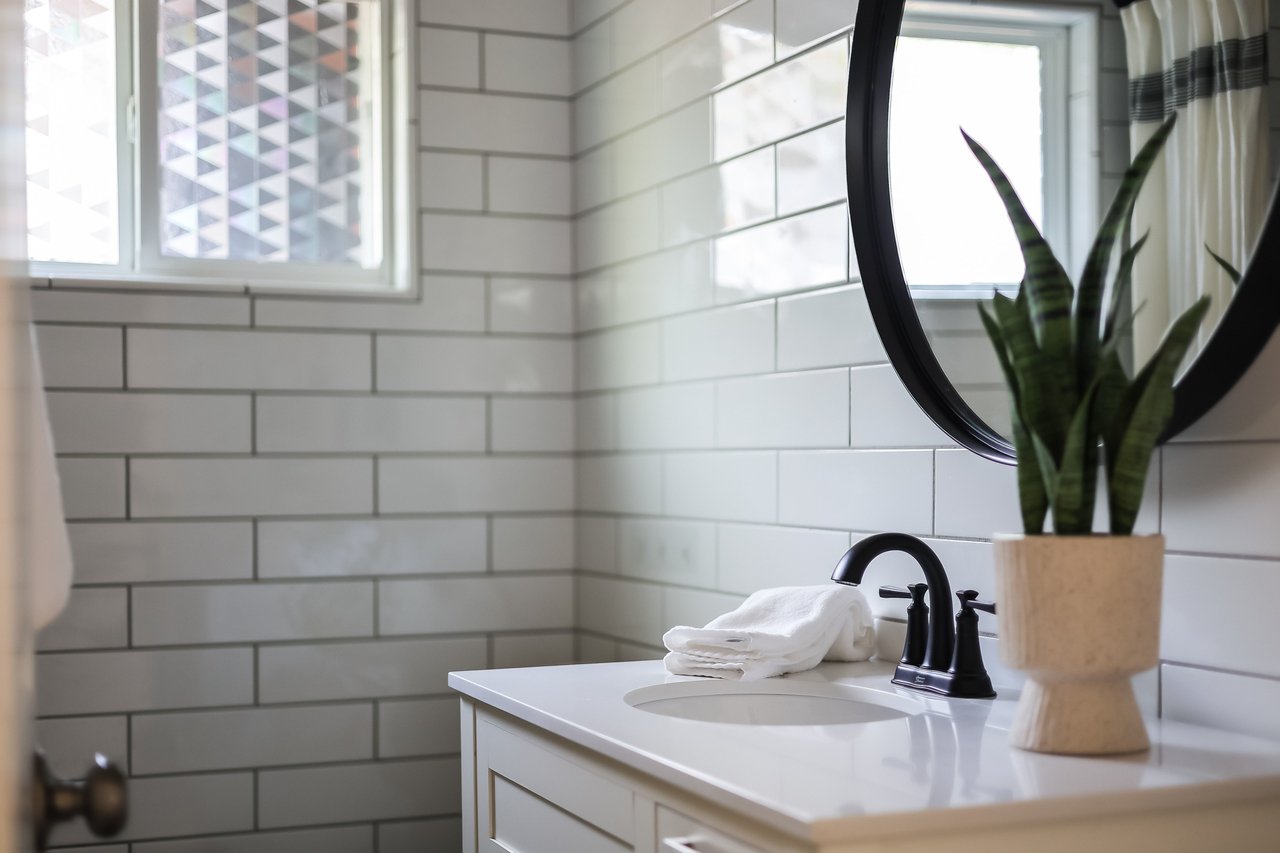 Bathroom vanity with white subway tile, a black faucet, round mirror, and potted plant.