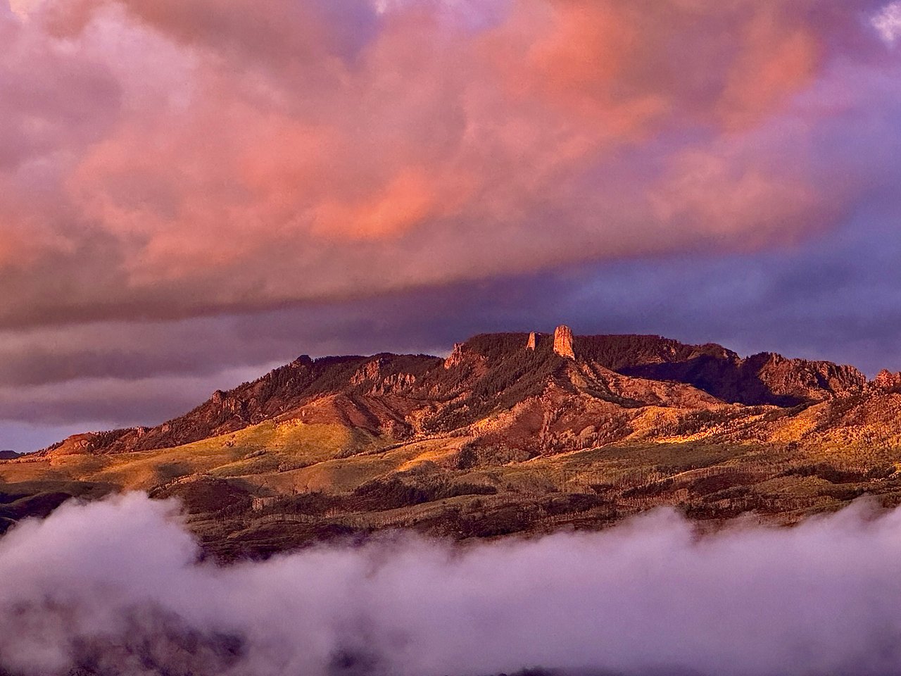 Rugged mesa and mountain ridges glow orange beneath pink clouds, with low clouds across the foreground.