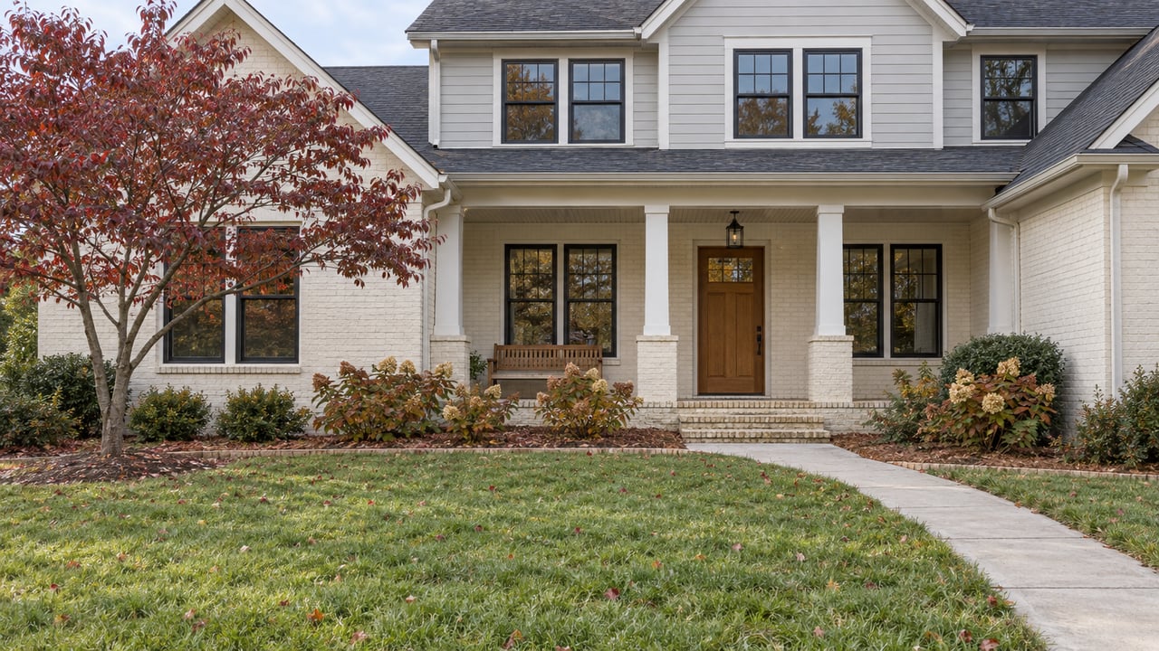 An ivory brick and pale gray sided home behind a deep lawn, with an oak front door, brick porch, and burgundy-leaved dogwood.