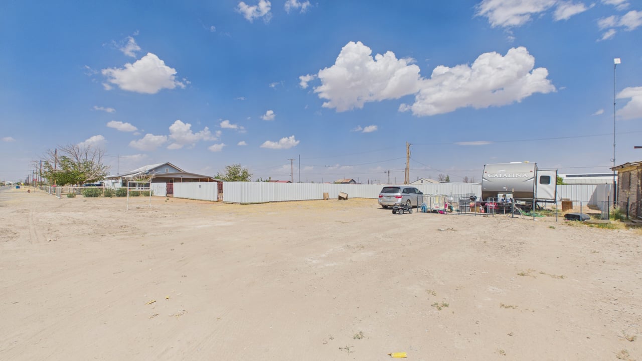 Fenced dirt lot with an RV and neighboring homes beneath a cloud-filled blue sky.