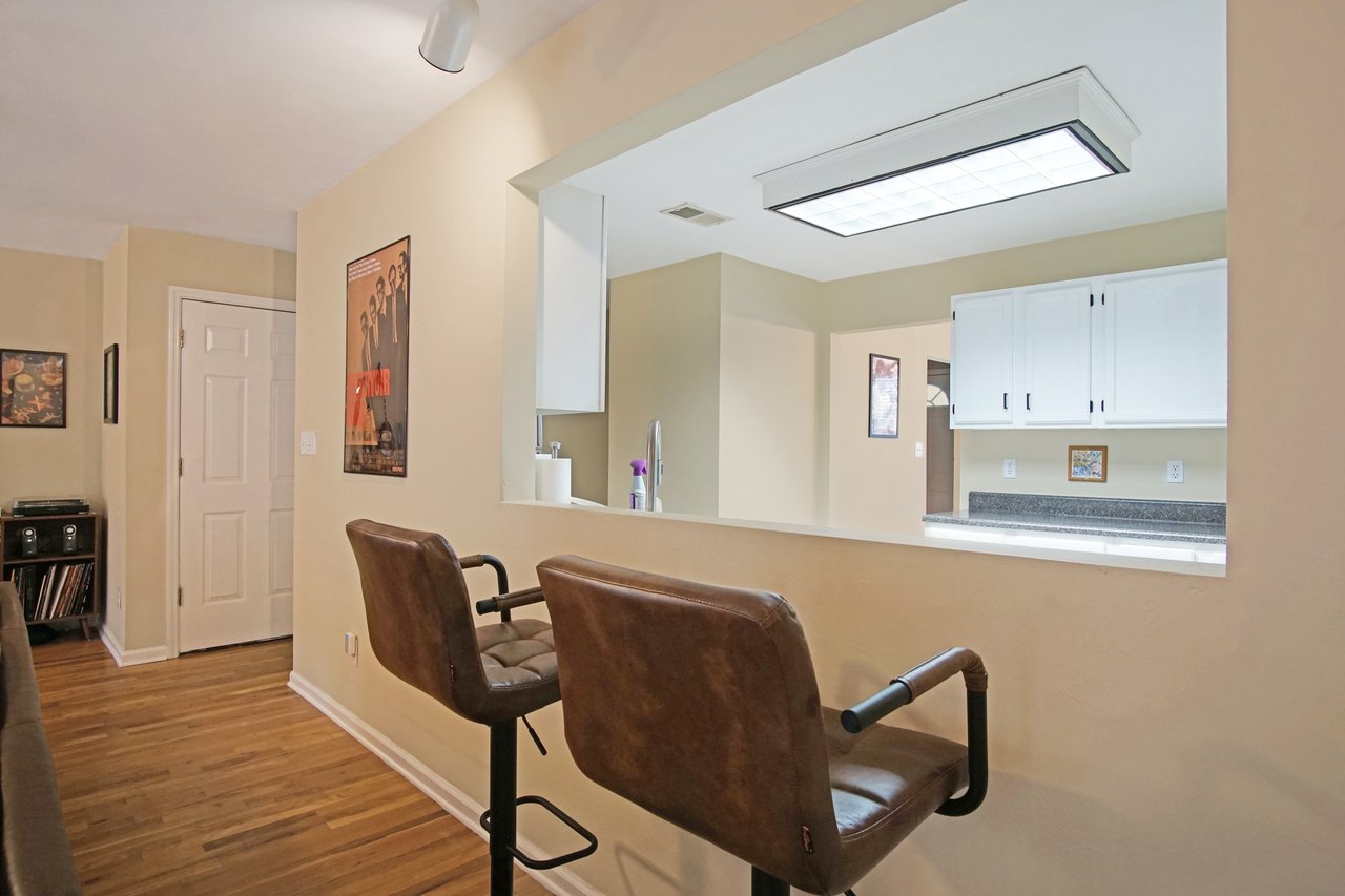 Kitchen pass-through and breakfast bar area at 529 Collinsford Road, Tallahassee, Florida, featuring two brown barstools, hardwood flooring, and beige walls. The view looks into the kitchen with white cabinetry, gray countertops, and a large ceiling light, creating an open and connected layout between living and dining spaces.