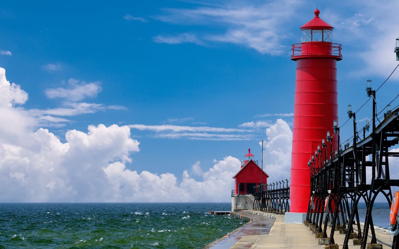 Red lighthouse on a long pier beside choppy water under a blue, cloud-filled sky.