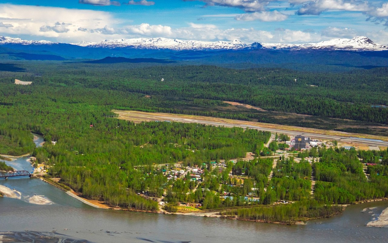 Aerial view of Talkeetna between a broad river, green forest, and snow-capped mountains.